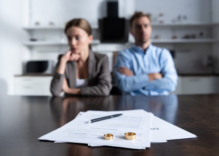 A close-up of official divorce petition paperwork and wedding rings on a wooden table, with an expat couple sitting blurred in the background, illustrating the legal divorce process in Mexico.