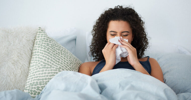 A sick woman in bed blowing her nose with a tissue, illustrating seasonal flu symptoms and the guide about flu shots in Merida.