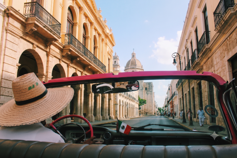 View from the backseat of a vintage convertible car driving down a sunny city street, illustrating practical road rules and transit safety guidelines for foreign drivers.